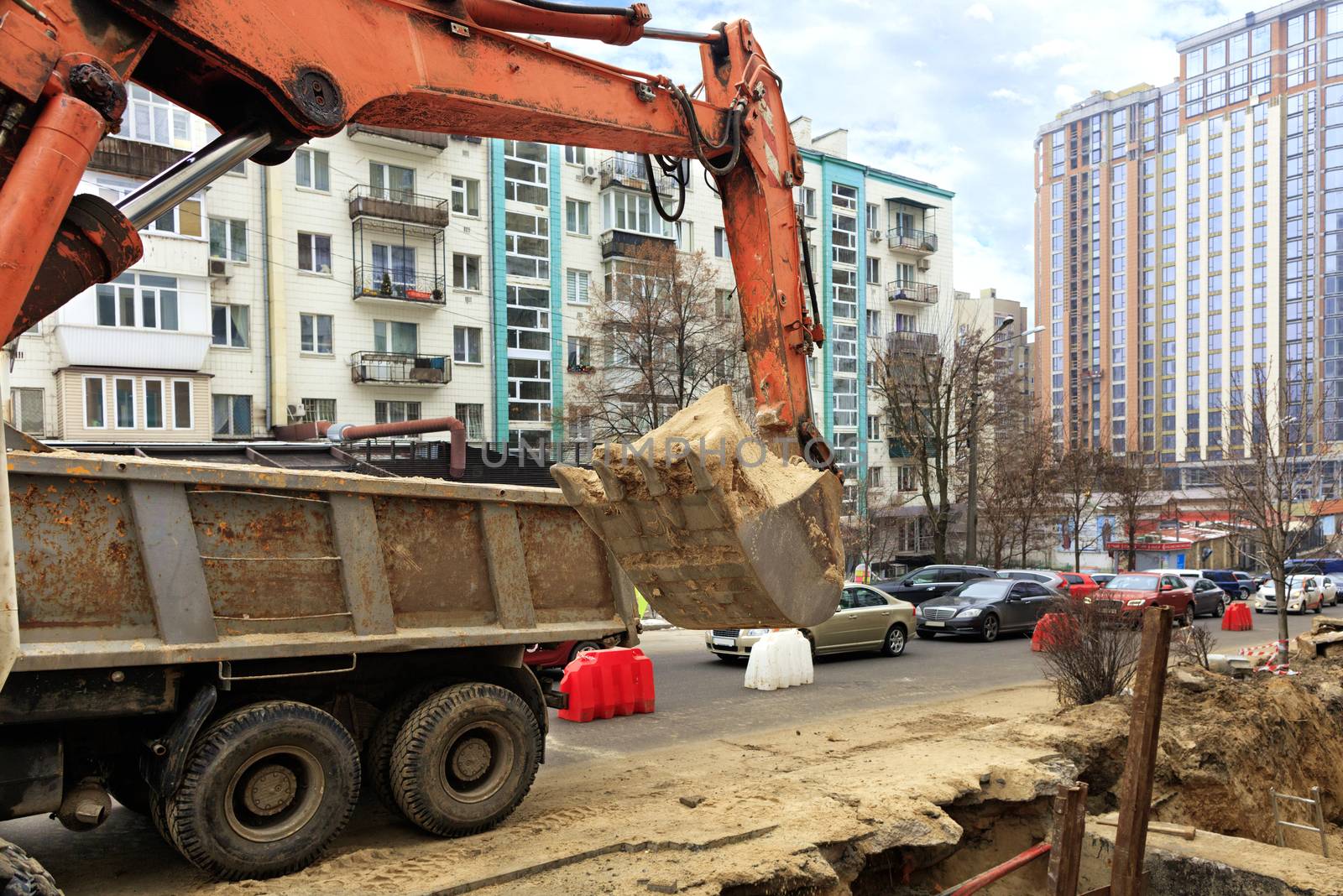 A heavy construction excavator bucket lifts sand into the back of a truck during a collector repair on a city street.