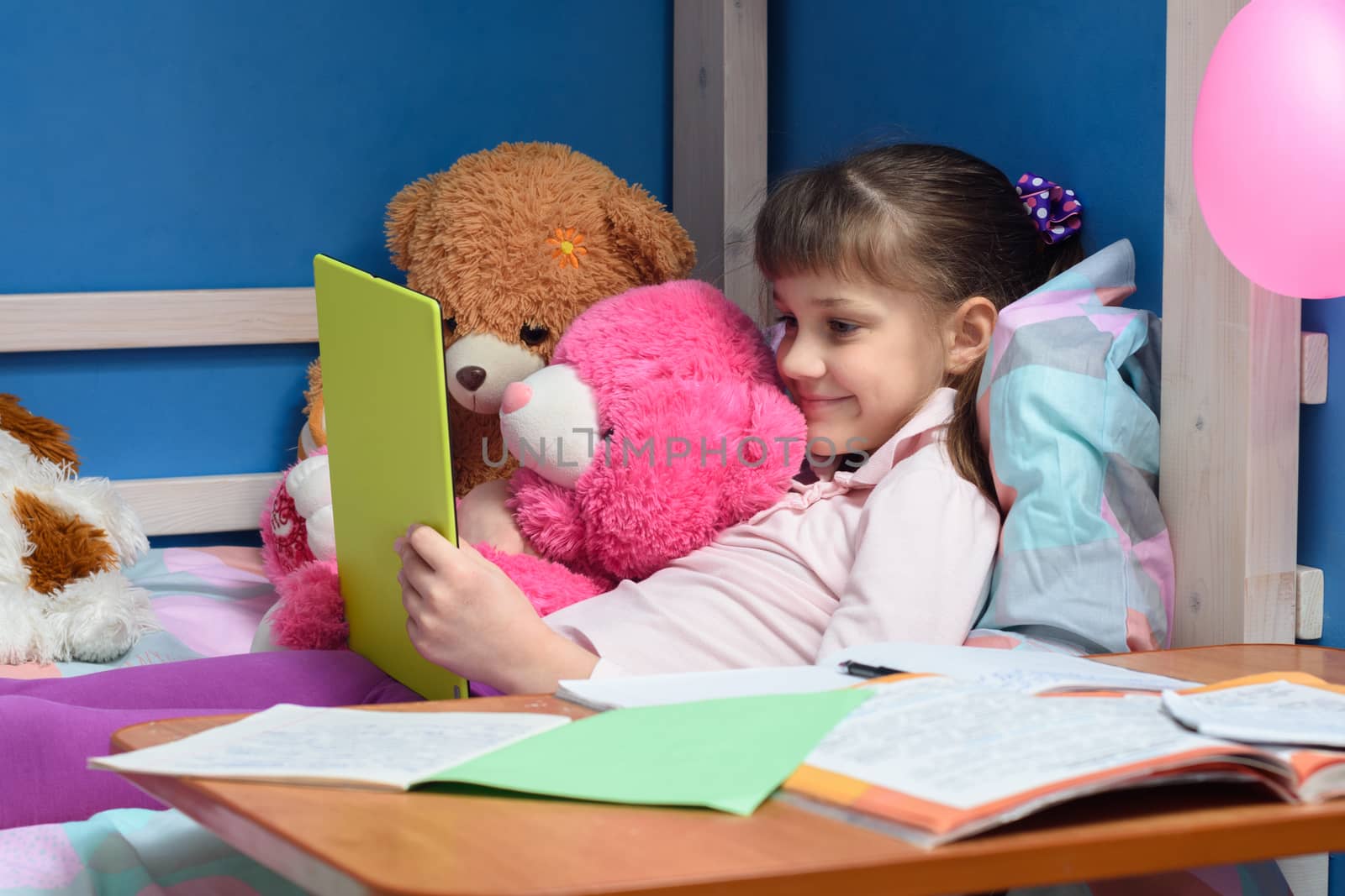 Nine-year-old girl lies on bed with toys and looks at tablet