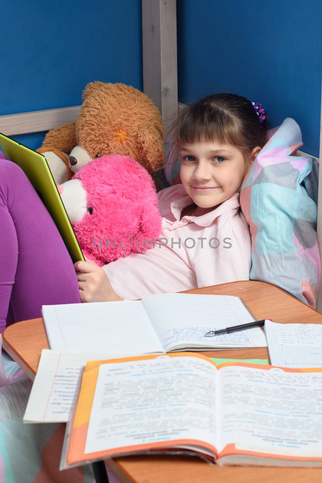 The girl looks into the frame with a smile holding a tablet, in front of her lie textbooks and notebooks