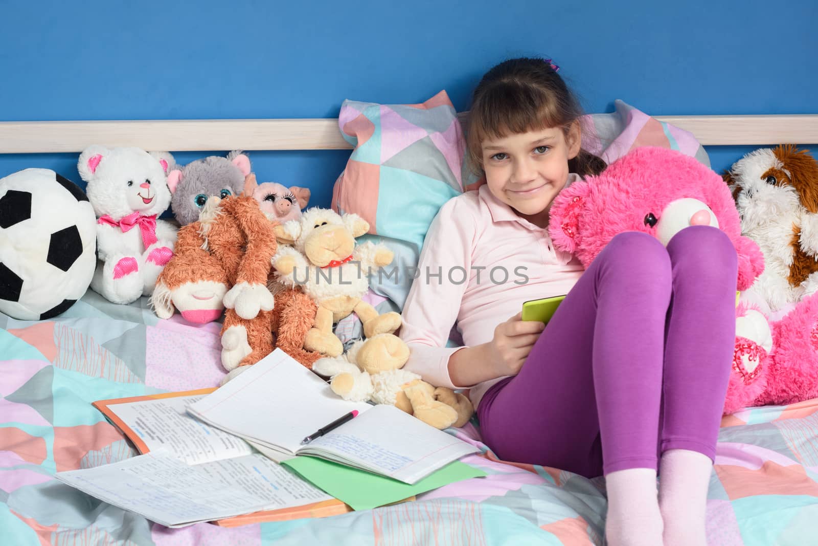 Girl among toys sits on the bed and does her homework