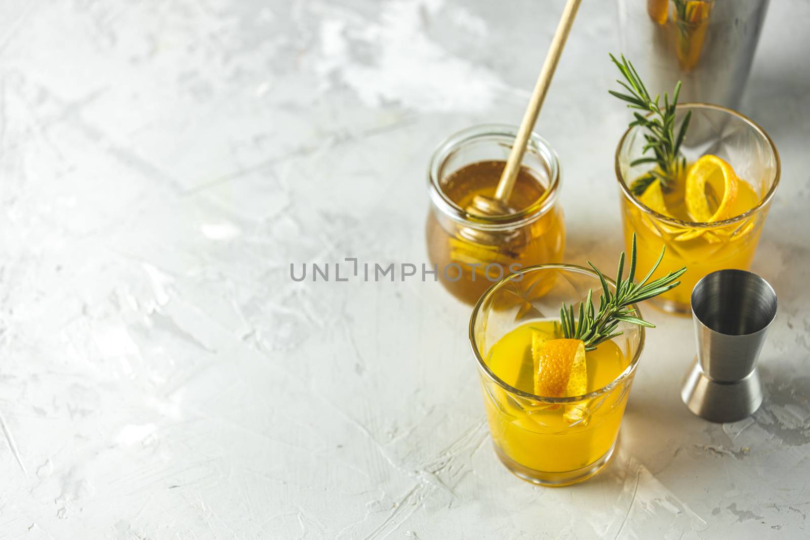 Two glasses of honey bourbon cocktail with rosemary simple syrup or homemade whiskey sour cocktail drink with orange and rosemary decoration orange peel, jar of honey and bartender tools