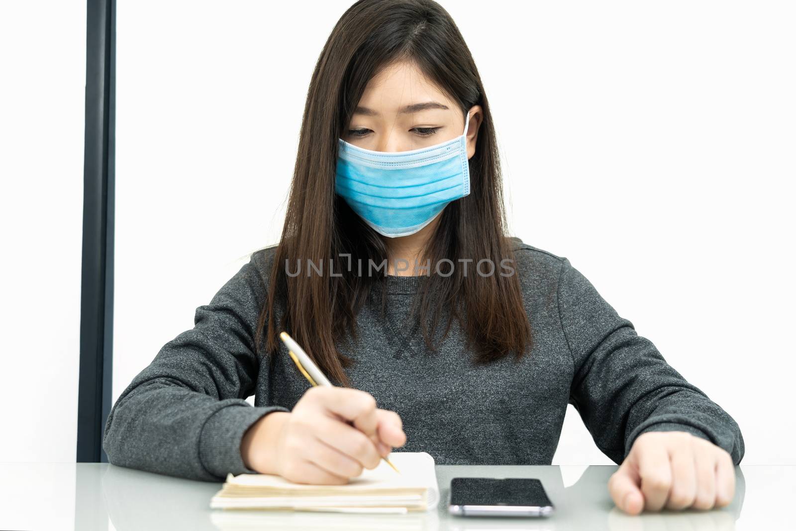 Teenage female student preparing for learnning at home sitting at a desk in front of smartphone with notepad and wear protective medical mask for protect Covid-19 or corona virus disease