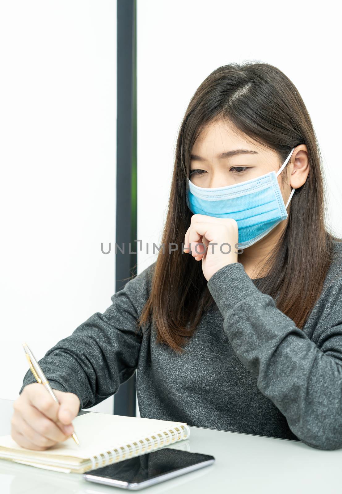 Teenage female student preparing for learnning at home sitting at a desk in front of smartphone with notepad and wear protective medical mask for protect Covid-19 or corona virus disease