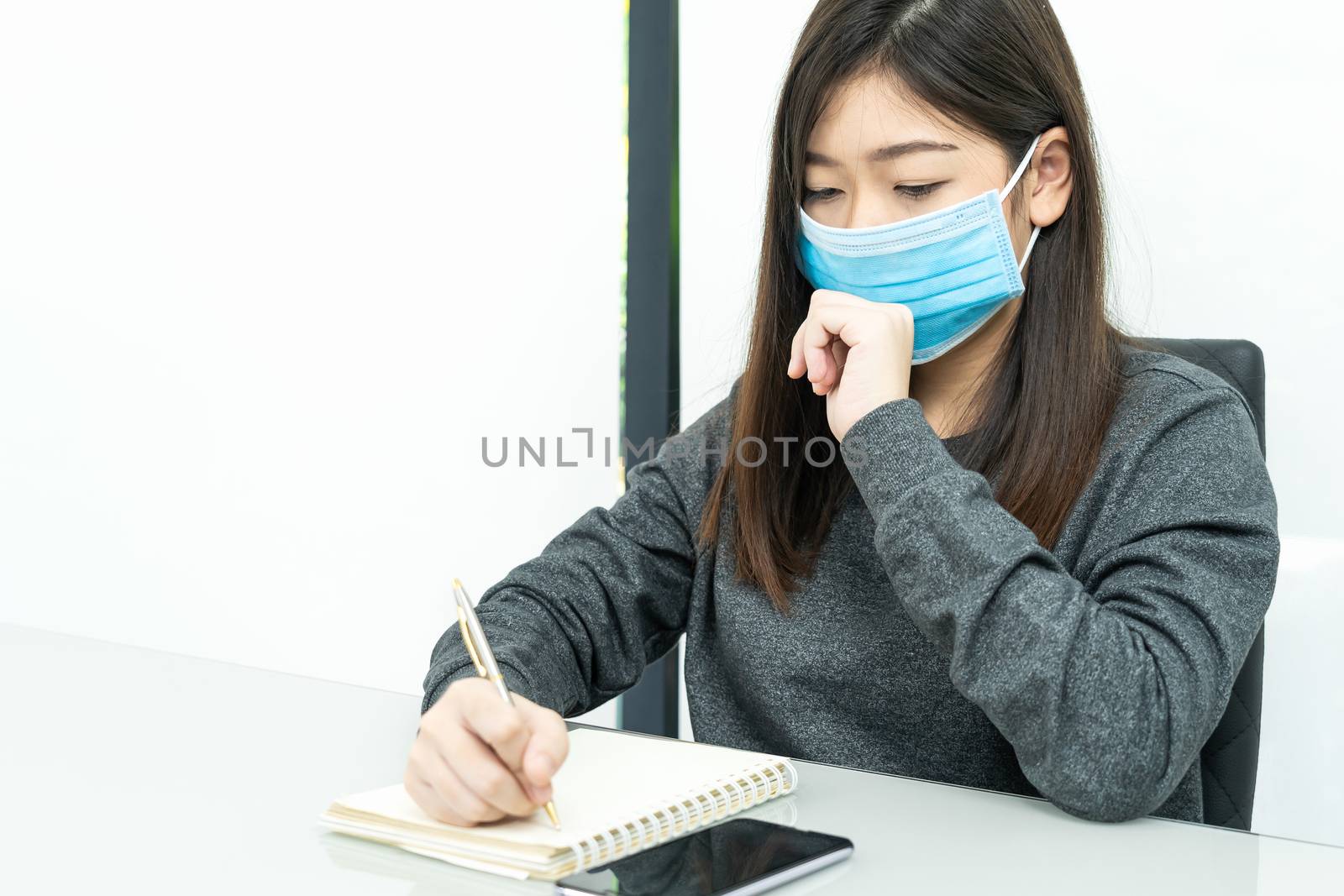 Teenage female student preparing for learnning at home sitting at a desk in front of smartphone with notepad and wear protective medical mask for protect Covid-19 or corona virus disease