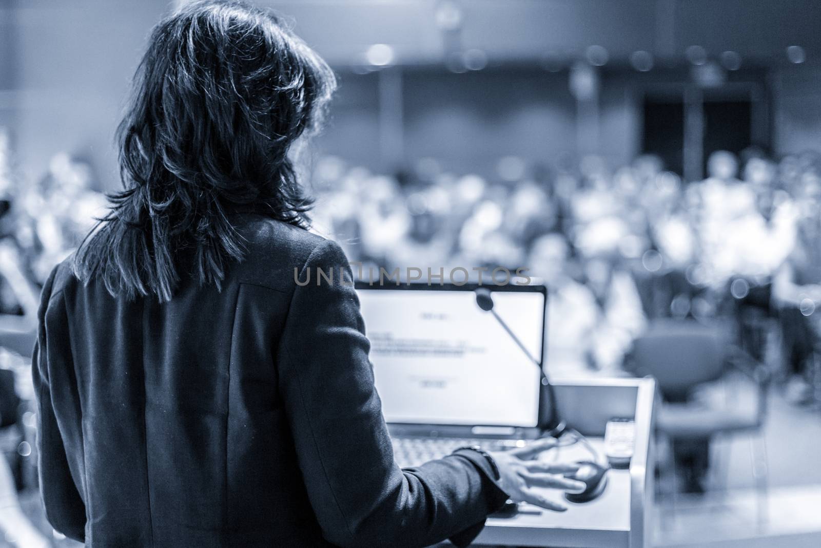 Female speaker giving a talk on corporate business conference. Unrecognizable people in audience at conference hall. Business and Entrepreneurship event. Black and white, blue toned image.