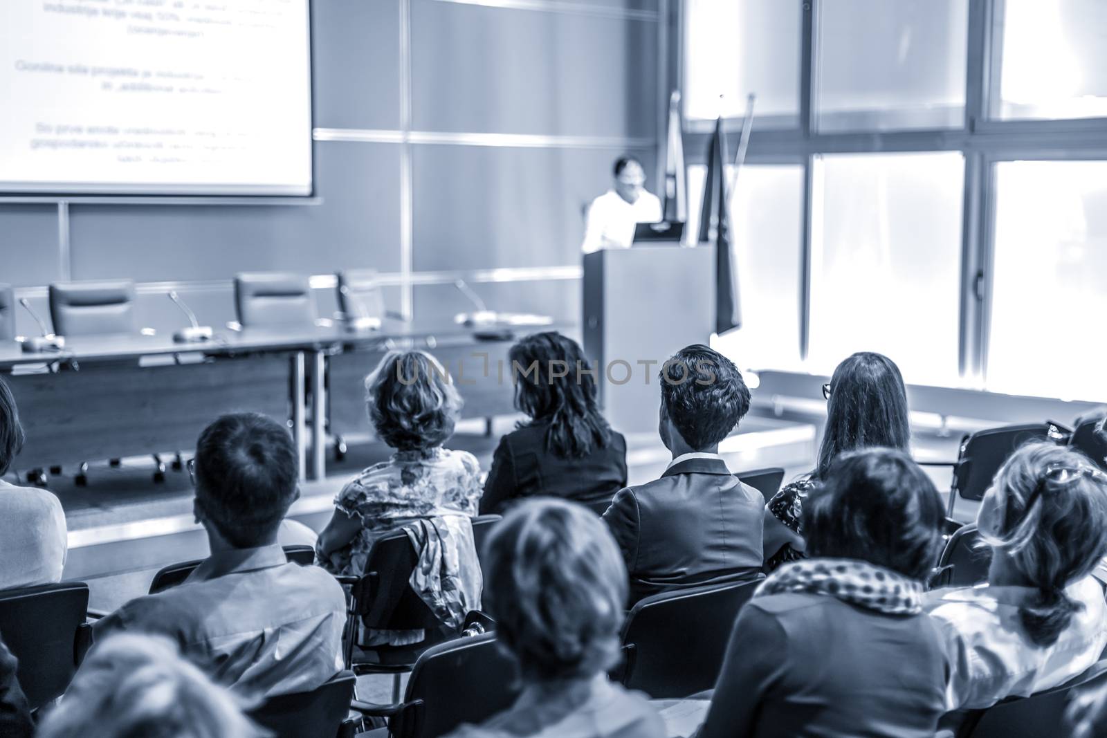 Female speaker giving presentation in lecture hall at university workshop. Audience in conference hall. Rear view of unrecognized participant in audience. Scientific conference event.