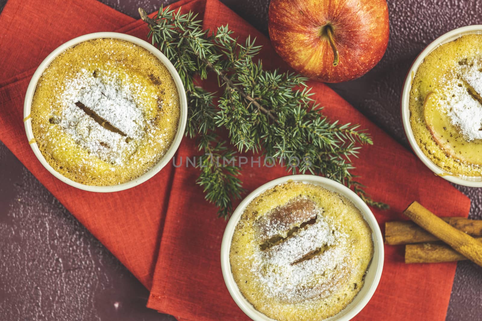 Three apple pies in ceramic baking molds ramekin with napkin, apple, spruce branch, cinnamon on dark red concrete table. Top view, flat lay. Christmas and New Year food background.