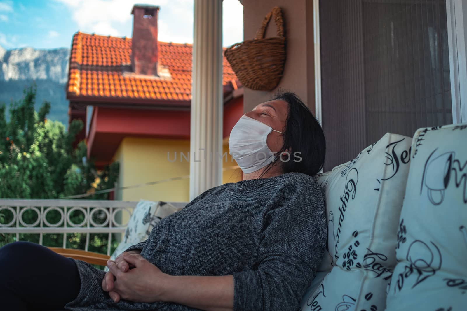 Middle aged caucasian woman with medical face mask, resting on cpuch on veranda during COVID-19 threat. by Emurado