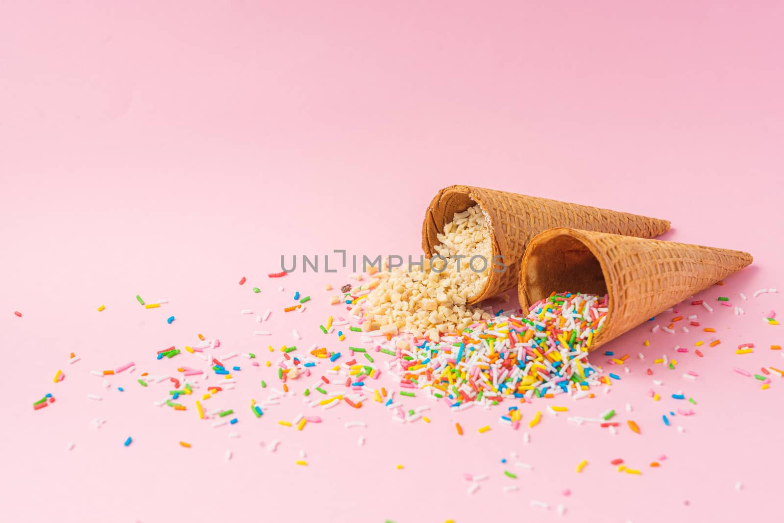 Two wheat flour ice cream cones one filled with roasted, natural and granulated almonds and the other filled with colored granulated sugar on a pink background. Summer concept