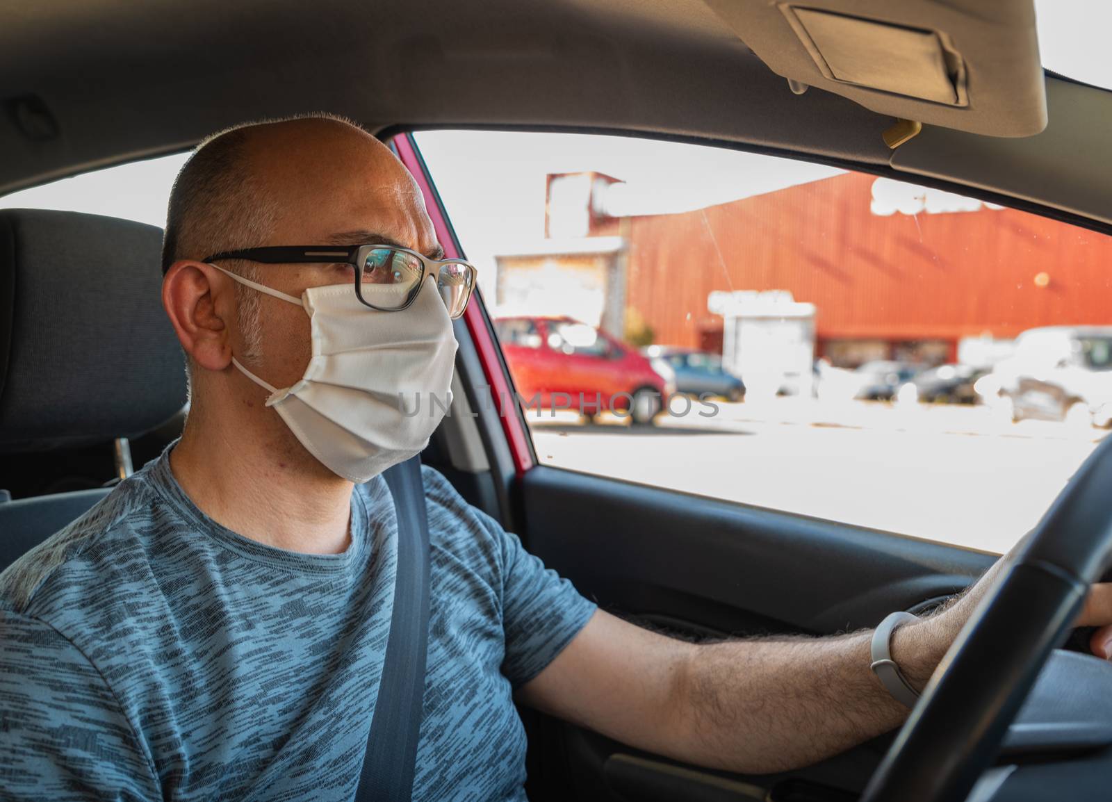 Turin, Piedmont, Italy. Maggio2020. Coronavirus pandemic: portrait of a Caucasian man driving a car wearing a white mask to avoid contagion. Selective focus on man and blurred background. Sunny day.