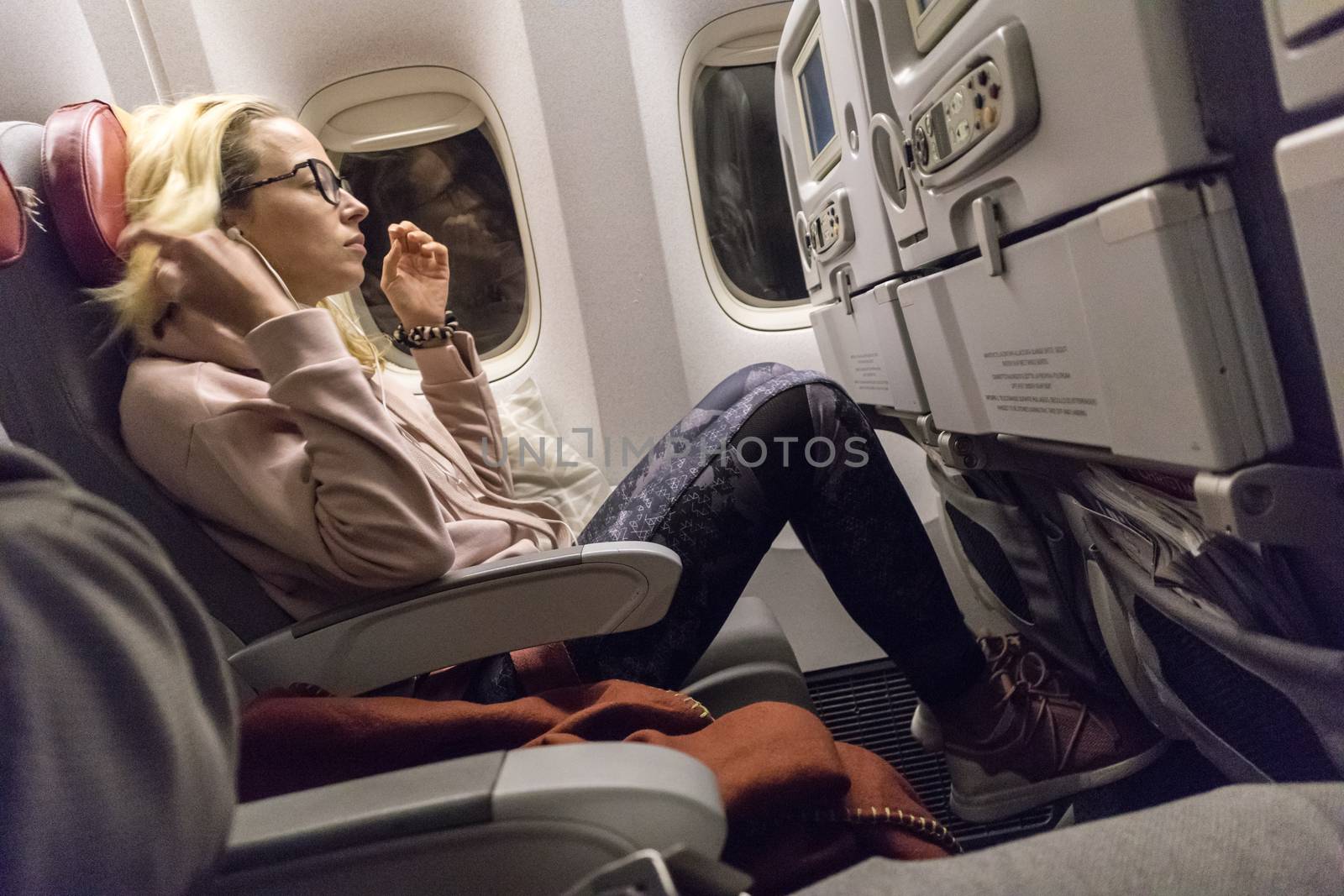 Sporty casual young blonde caucasian lady watching movie while traveling by airplane by the window. Commercial transportation by planes.