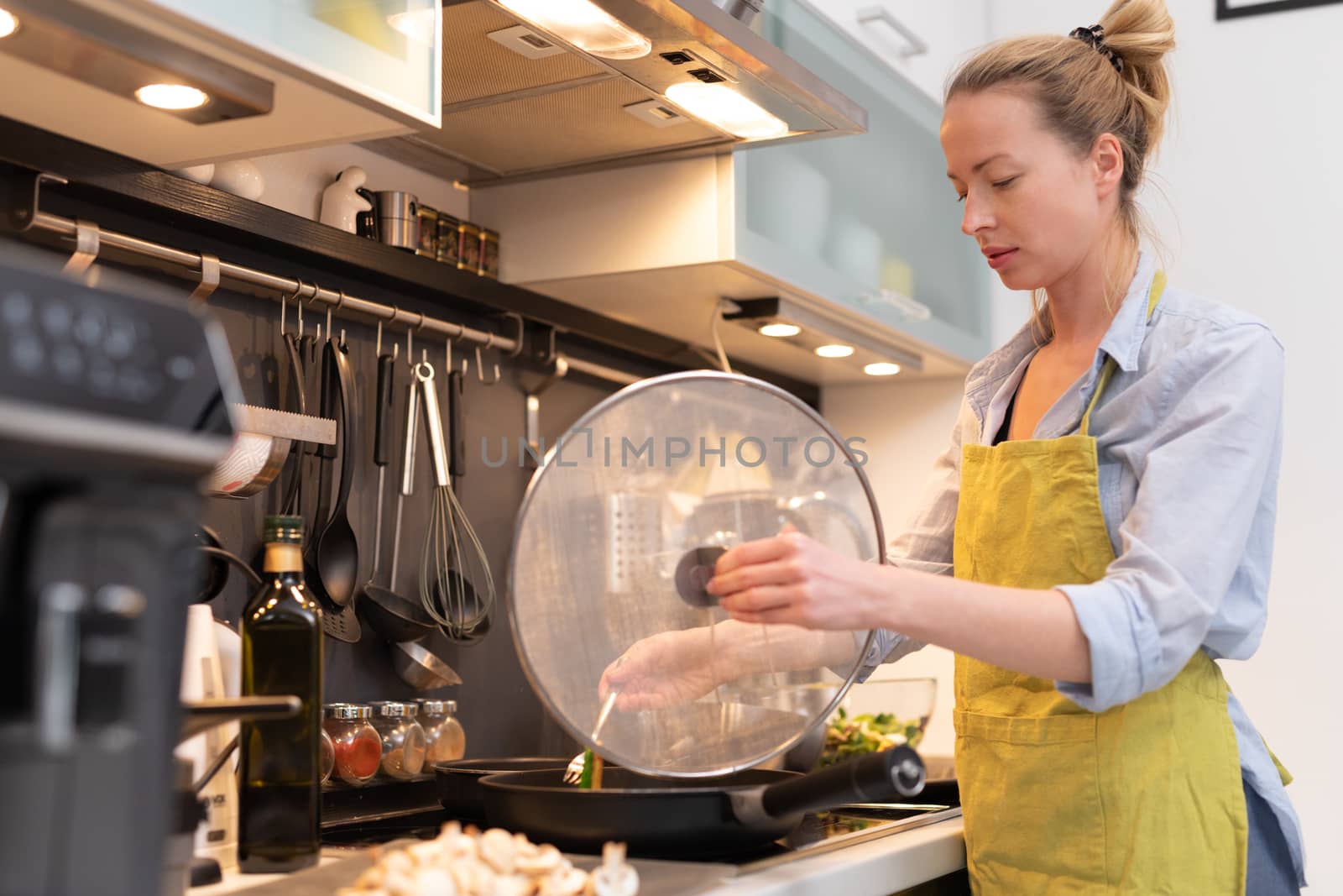 Stay at home housewife woman cooking in kitchen, stir frying dish in a saucepan, preparing food for family dinner. Blonde caucasian model in her 30is, real cooking not looking in camera.