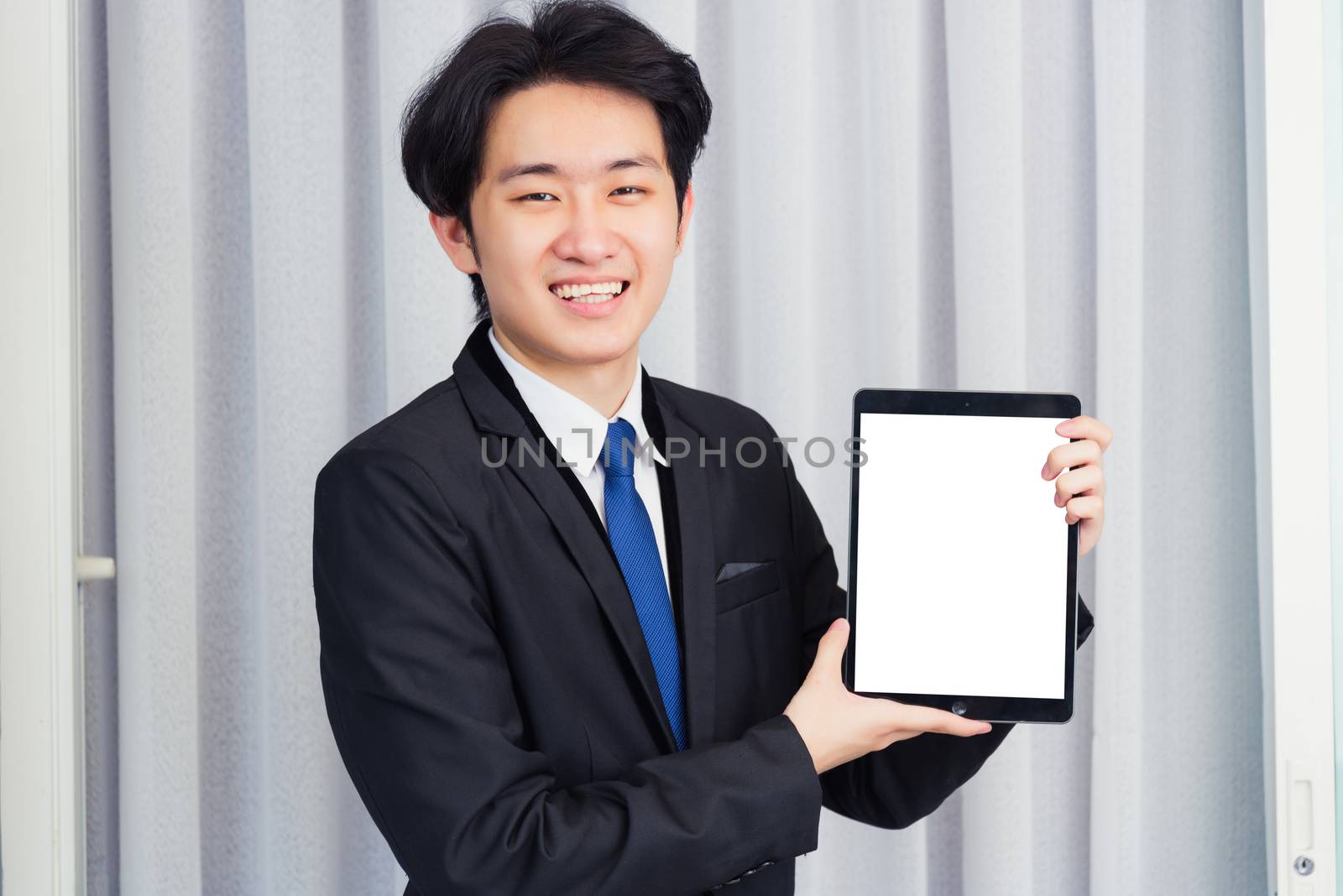 Work from home, Asian young businessman wear suit video conference call or facetime he smiling and showing blcnk screen of smart digital tablet computer at home office, looking to camera