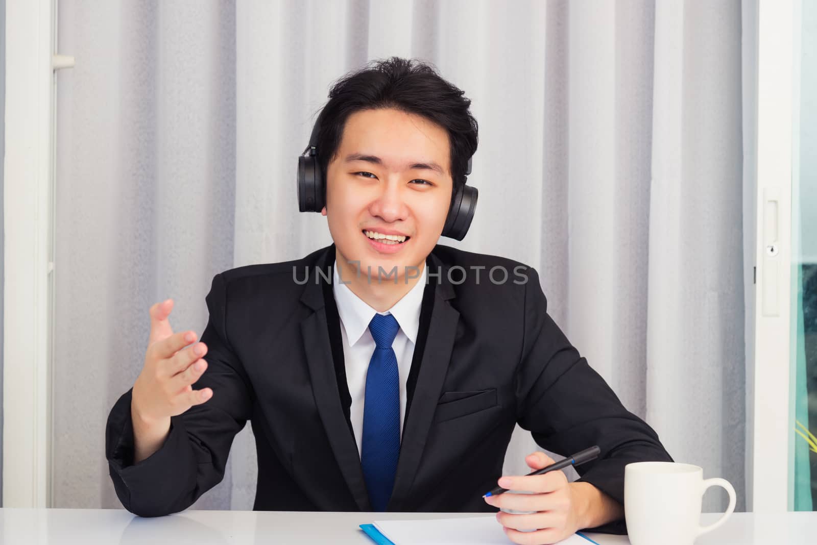 Work from home, Asian young businessman wear suit video conference call or facetime he smiling sitting on desk wearing headphones and raise his hand to explain the job he looking to camera