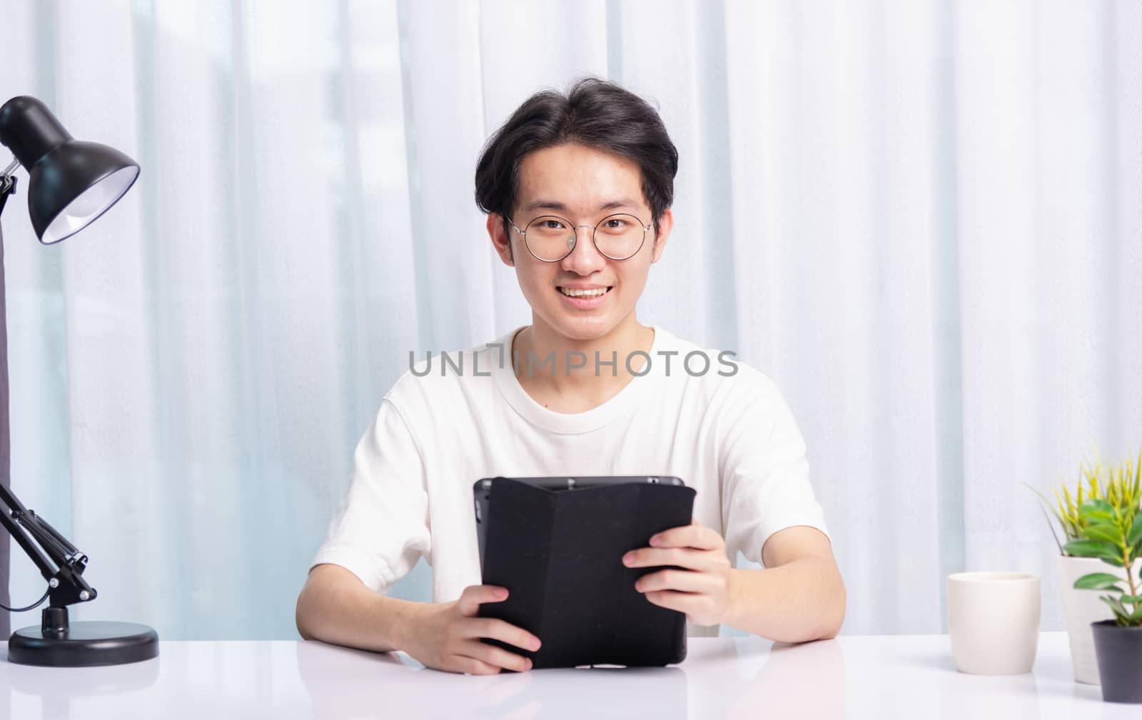 Happy Asian young business handsome man work from home office wear glasses, t-shirt comfortable he smiling and using a black modern smart digital tablet computer to read email or learning on desk