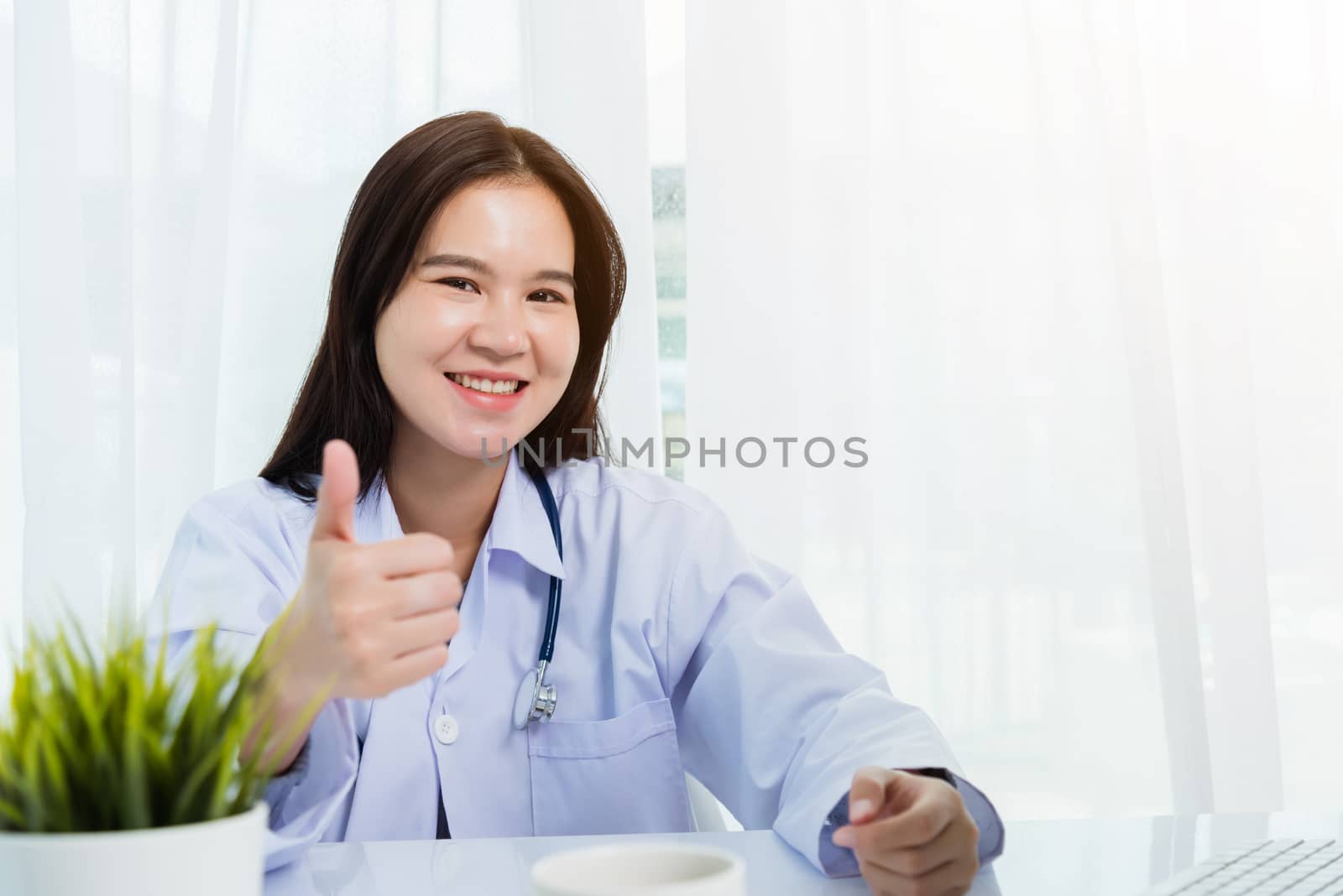 Asian young beautiful woman doctor raise hand finger thumb up for good sign on front desktop PC computer monitor quarantines disease coronavirus or COVID-19 at hospital office