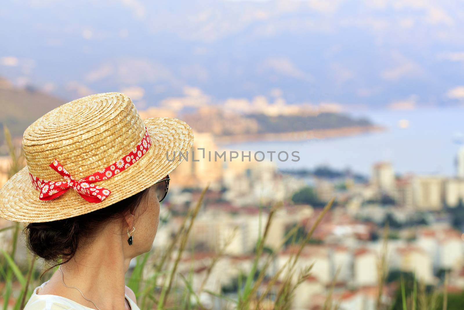 A young woman in a straw hat with a motley red ribbon and in sunglasses is looking from a height on the bay and the city of Budva in blur, Montenegro.