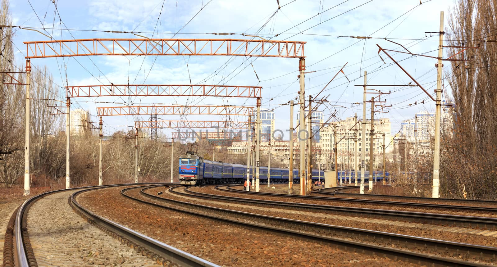 Passenger train cars of the train ride on the railway tracks in the background of the cityscape by Sergii