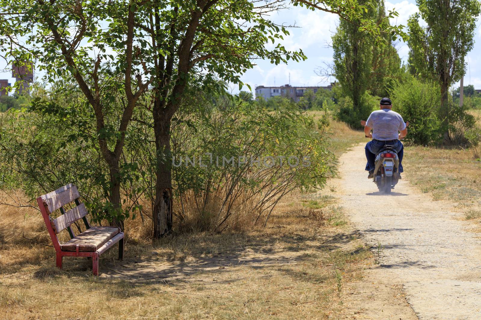 A bench in the shade of a tree, old and weathered, against the backdrop of a rural landscape and a resident who is riding a scooter along an old concrete path.