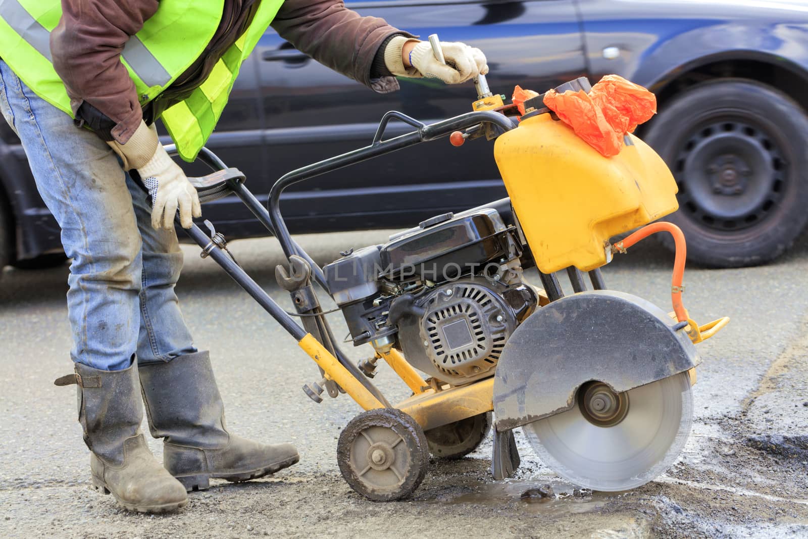 An employee of a road maintenance service in a green reflective vest removes old asphalt with a gasoline cutter during repairs on the roadway.
