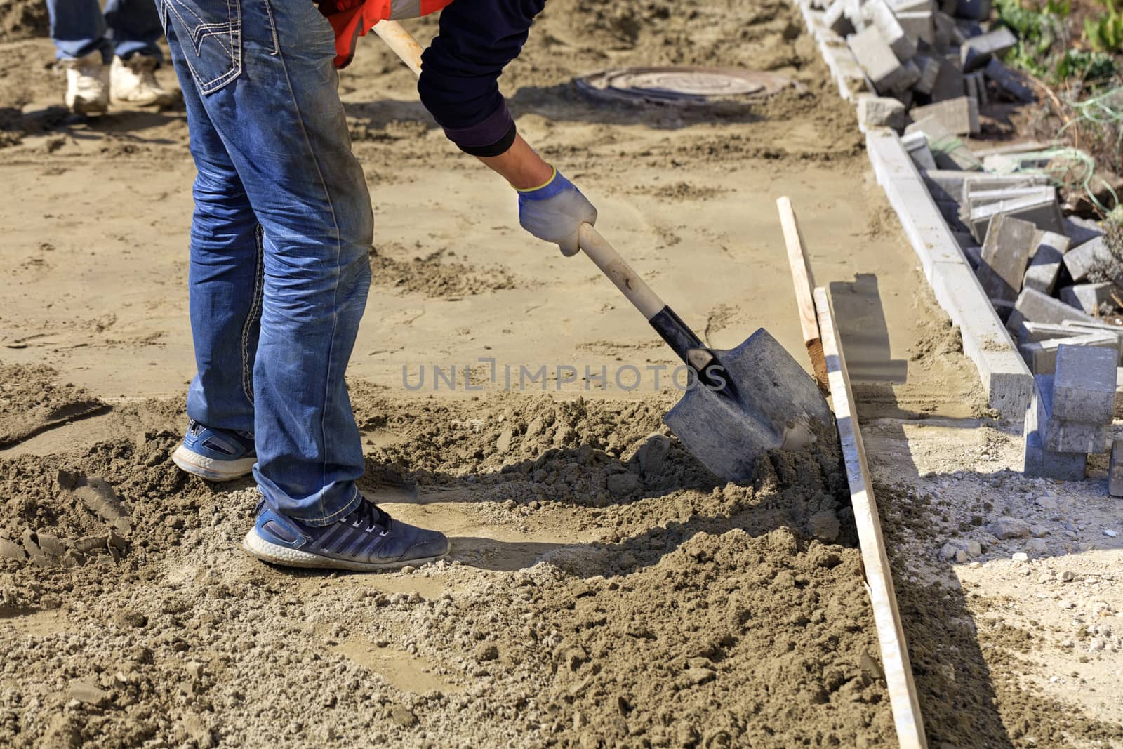 The worker aligns the foundation with sand and a shovel under the wooden level for laying paving slabs on the background of the site prepared for work.