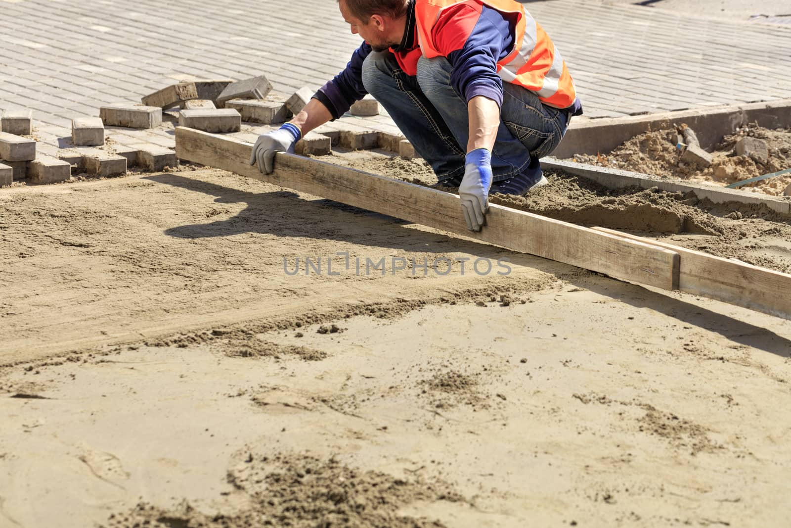 On the sidewalk, a worker aligns the sand platform with a wooden board, preparing the foundation for laying paving slabs.