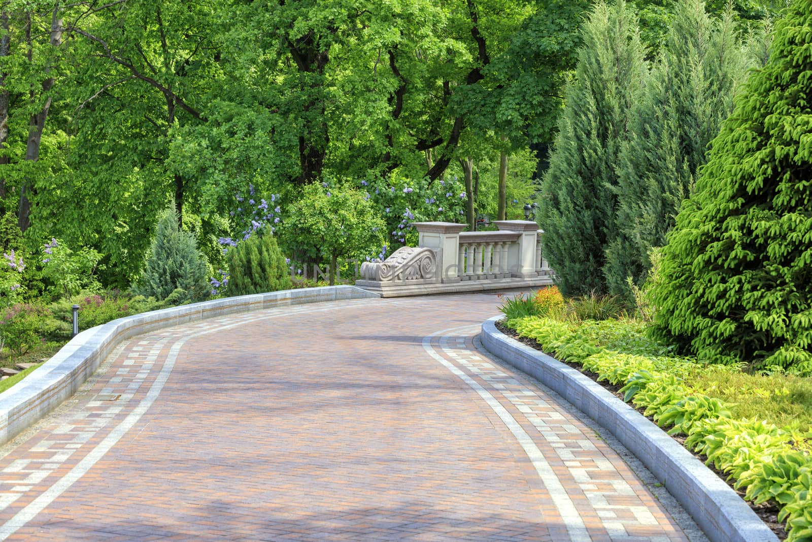 A laid flat path in a beautiful park passes through a green lawn with decorative flowers, framed on both sides with various bushes, well-kept trees and lanterns under soft sunlight.