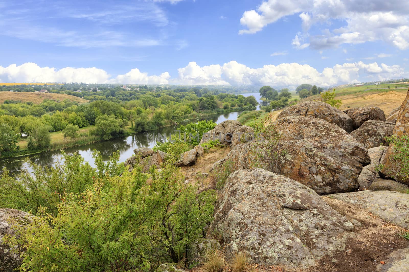 The Southern Bug River in the summer is rocky shores, bright green vegetation and a cloudy blue sky.