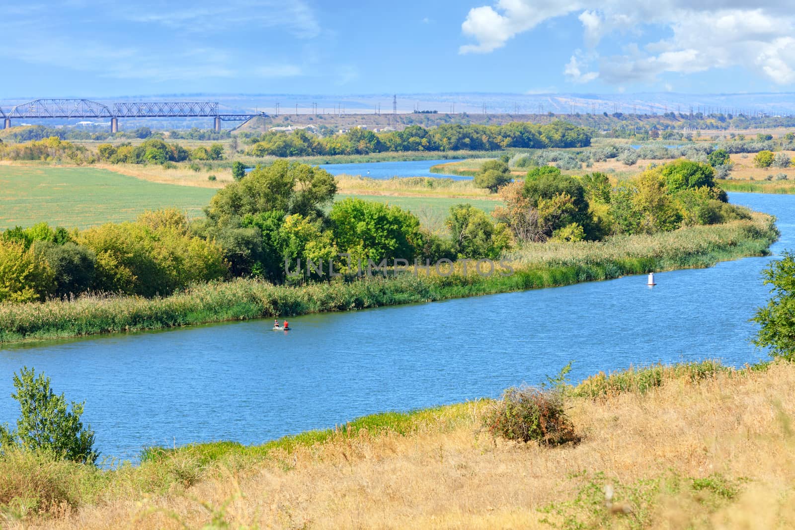 The wide bend of the Southern Bug River is immersed in the greenery of the garden and dried grass on the slope in the foreground, the river reaches the railway bridge on the horizon.