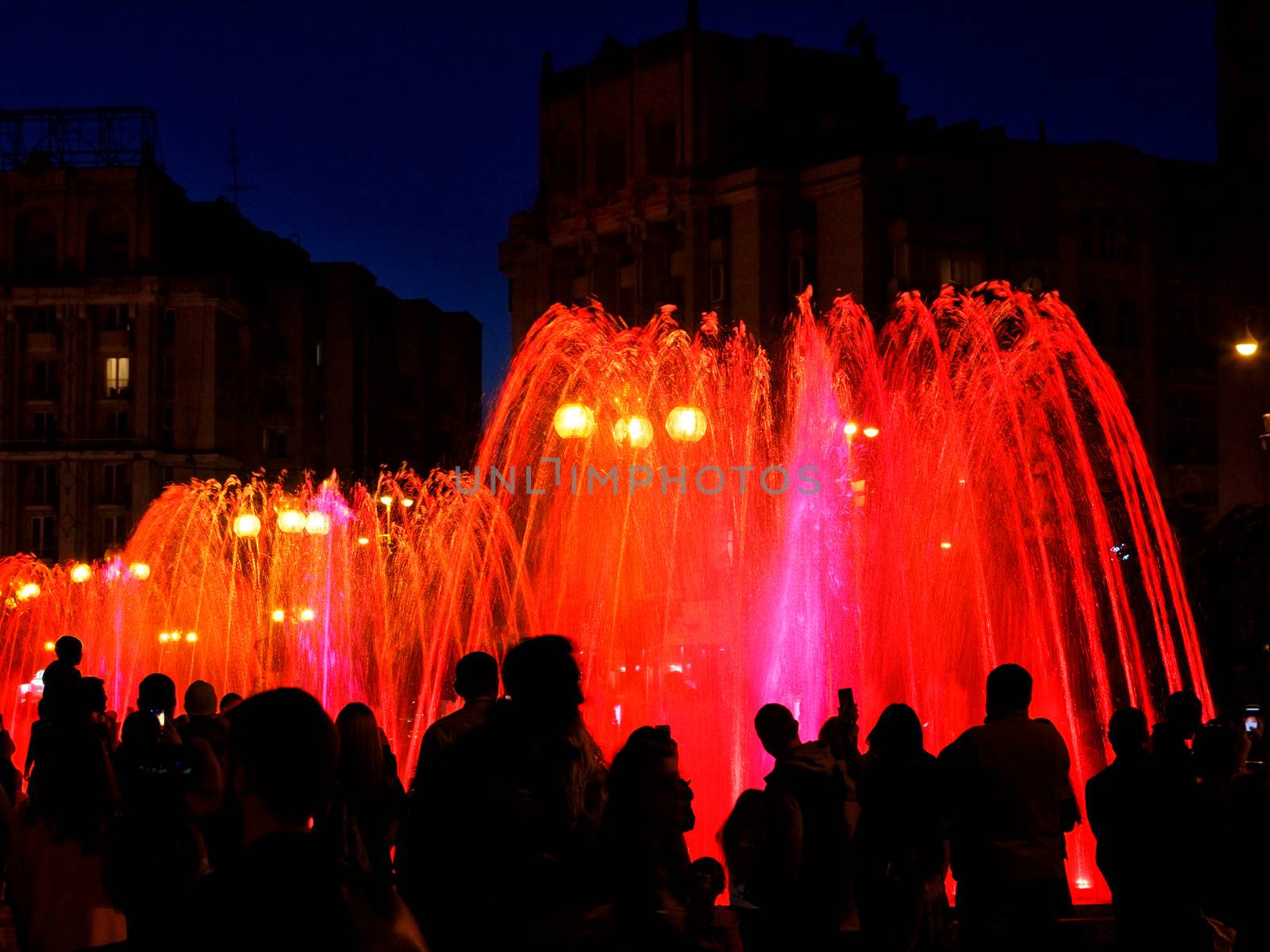Dark silhouettes of people around the fountainin the late evening, red neon lights illuminate the jets of a powerful urban singing fountain.