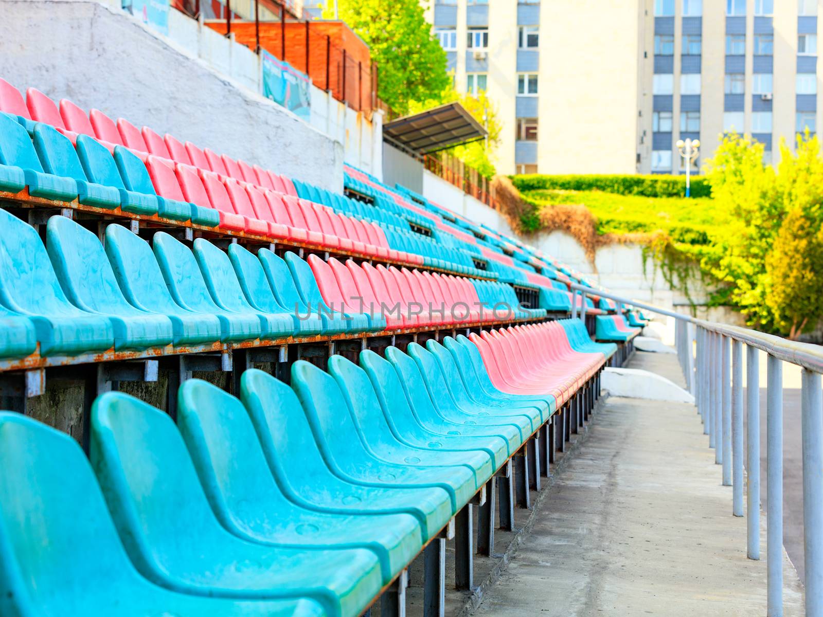 The rhythmic rows of empty turquoise and red plastic chairs of an old sports stadium against the background of sunlight in the background.