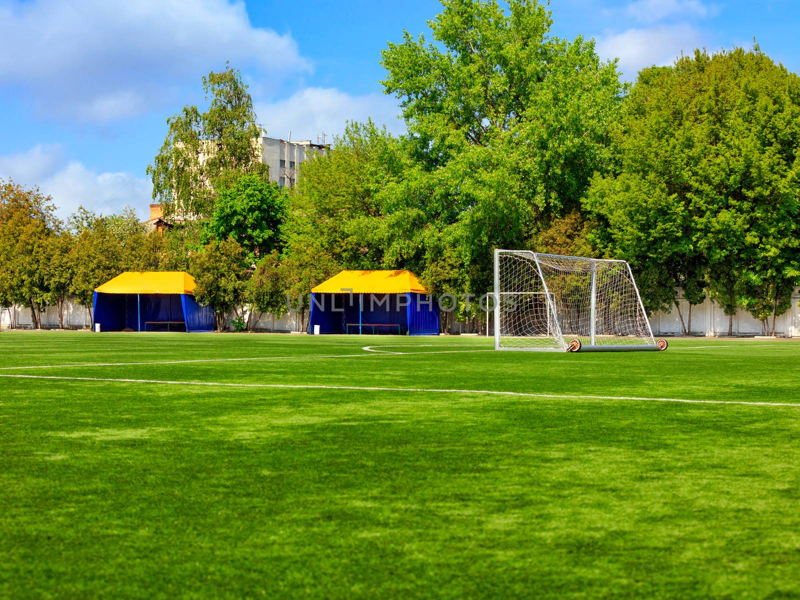 Green lawn of a football field with gates, netting and tents for team players on a summer day against a background of green foliage and a slightly cloudy blue sky.