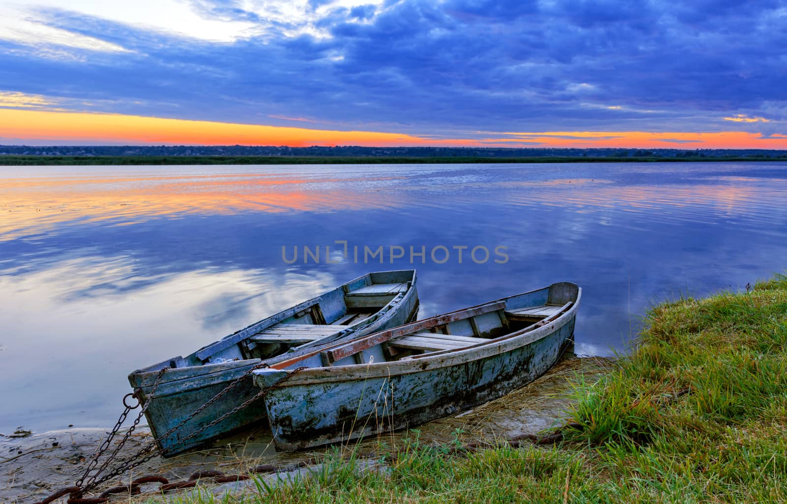 Two old blue-green wooden boats chained by the shore of a calm river. Early morning, the rays of the rising sun illuminate the summer landscape of the shore of a wide river.