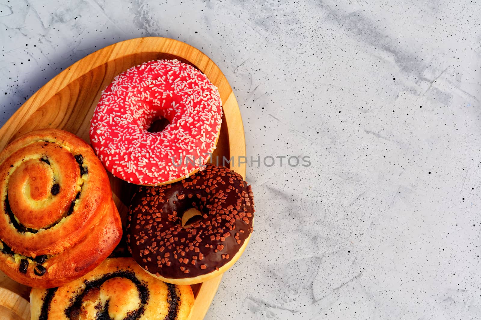Delicious donuts with chocolate and strawberry icing and buns with poppy seeds and freshly baked raisins, close-up on an oval wooden tray and a gray concrete surface. Pastry sweets for breakfast, image with copy space.