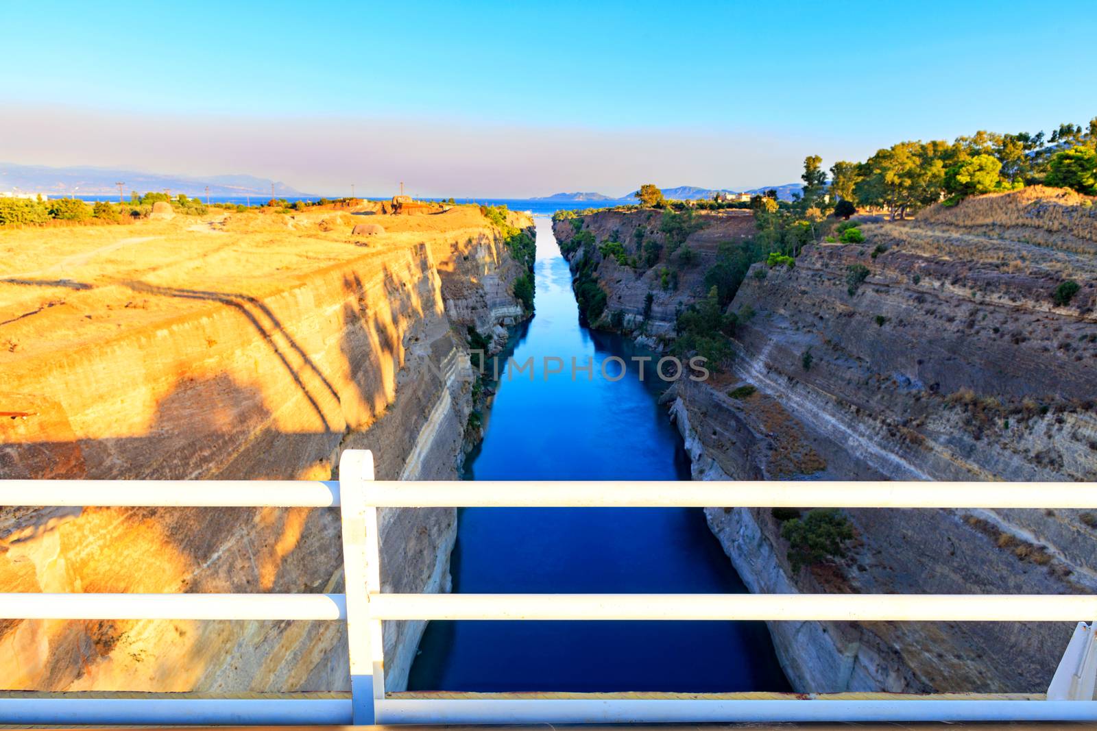 The Corinth Canal on a morning summer day illuminates the bright rising sun of Greece, a view of the Gulf of Corinth from the height of a pedestrian bridge.