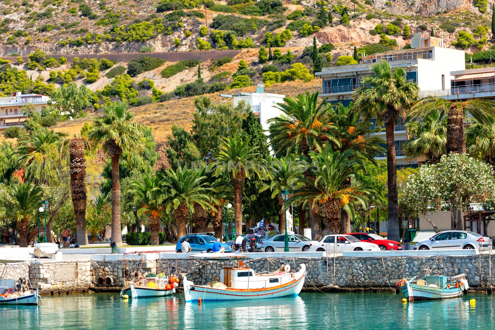 Old fishing schooners and boats moored at the pier of the Gulf of the Ionian Sea against the backdrop of the palm grove of the city park and the mountainside of Loutraki, Greece.