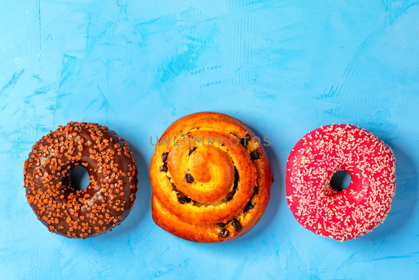 Delicious donuts with chocolate and strawberry icing and buns with and freshly baked raisins, sprinkled with coconut and fruit chips on top, close-up on a light turquoise surface. Pastry sweets for breakfast, image with copy space.