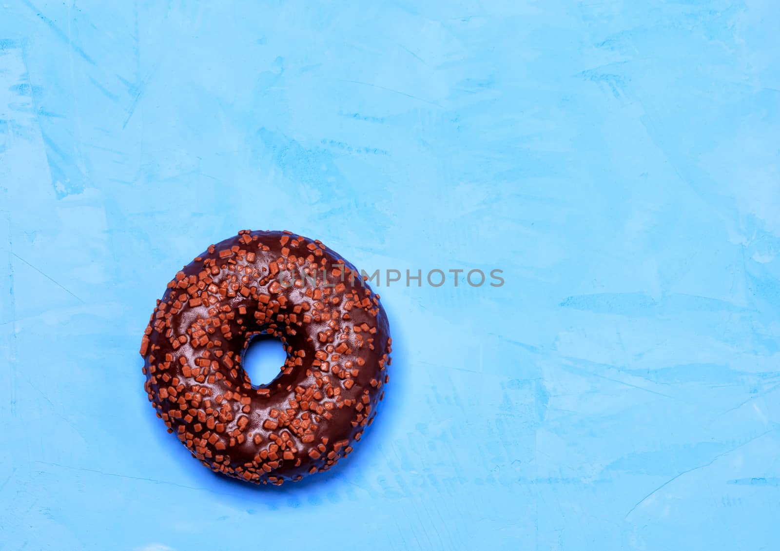 A delicious donut with chocolate icing sprinkled with fruit chips on top, close-up on a blue concrete surface with cold light. Pastry sweets for breakfast, image with copy space.