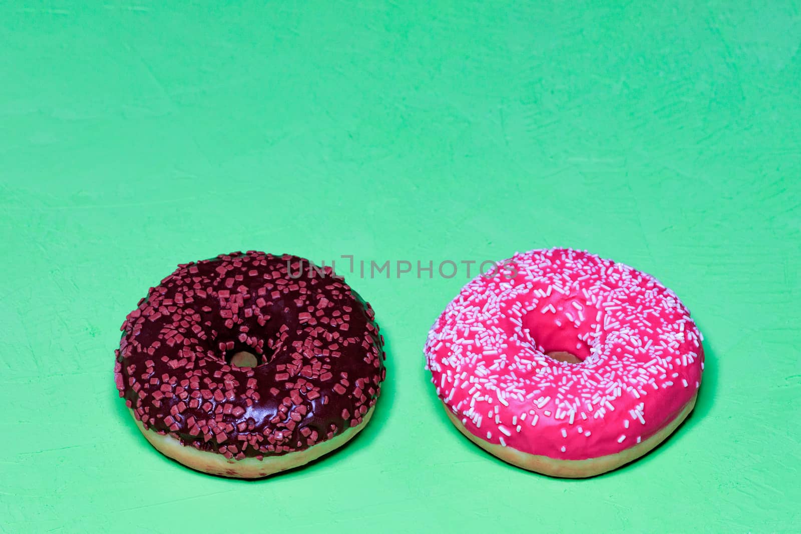 Two delicious donuts with chocolate and strawberry icing sprinkled with coconut and fruit chips on top, close-up on a light green rough surface. Pastry sweets for breakfast, image with copy space.