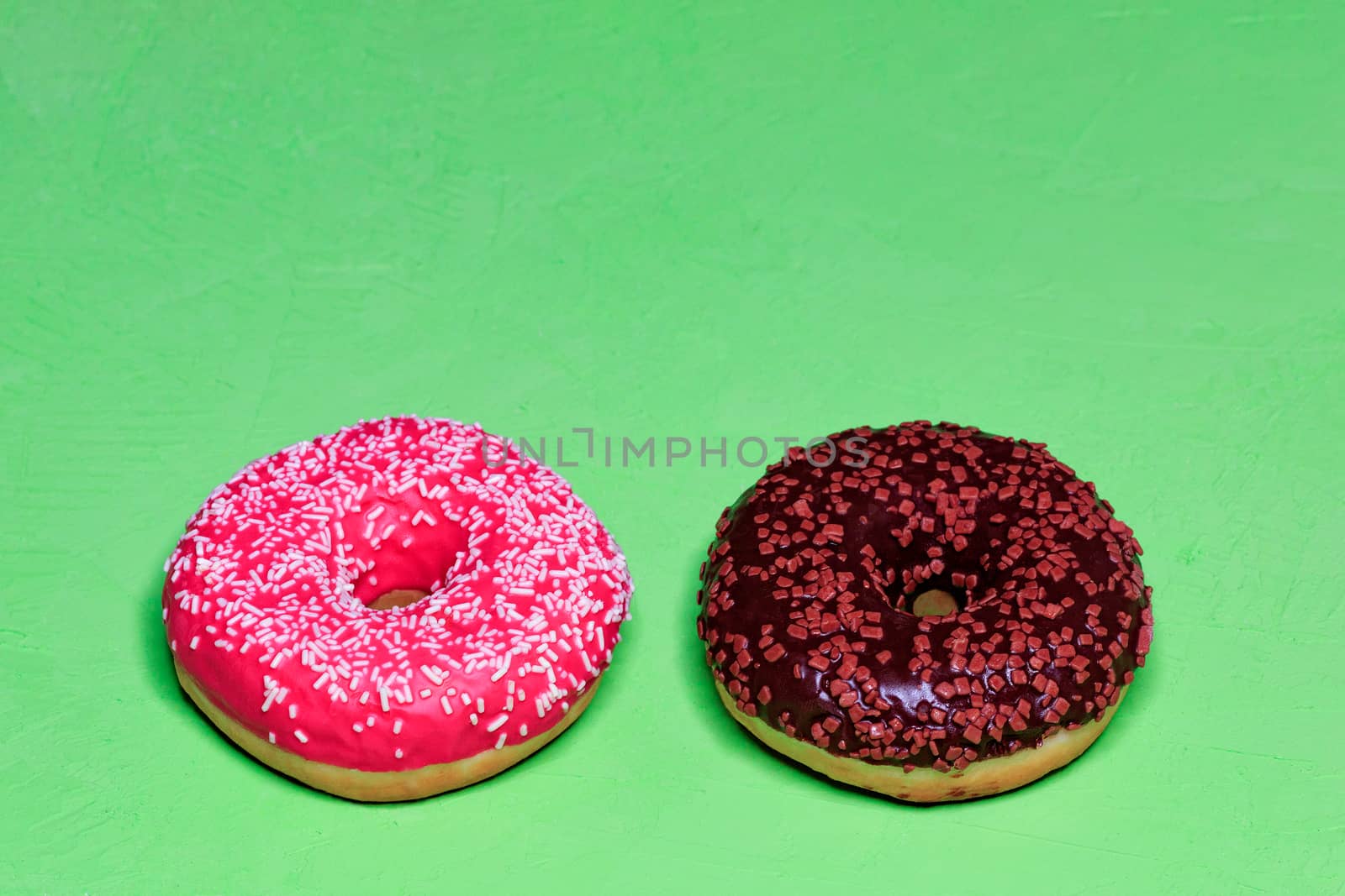 Two delicious donuts with chocolate and strawberry icing sprinkled with coconut and fruit chips on top, close-up on a green rough surface. Pastry sweets for breakfast, image with copy space.