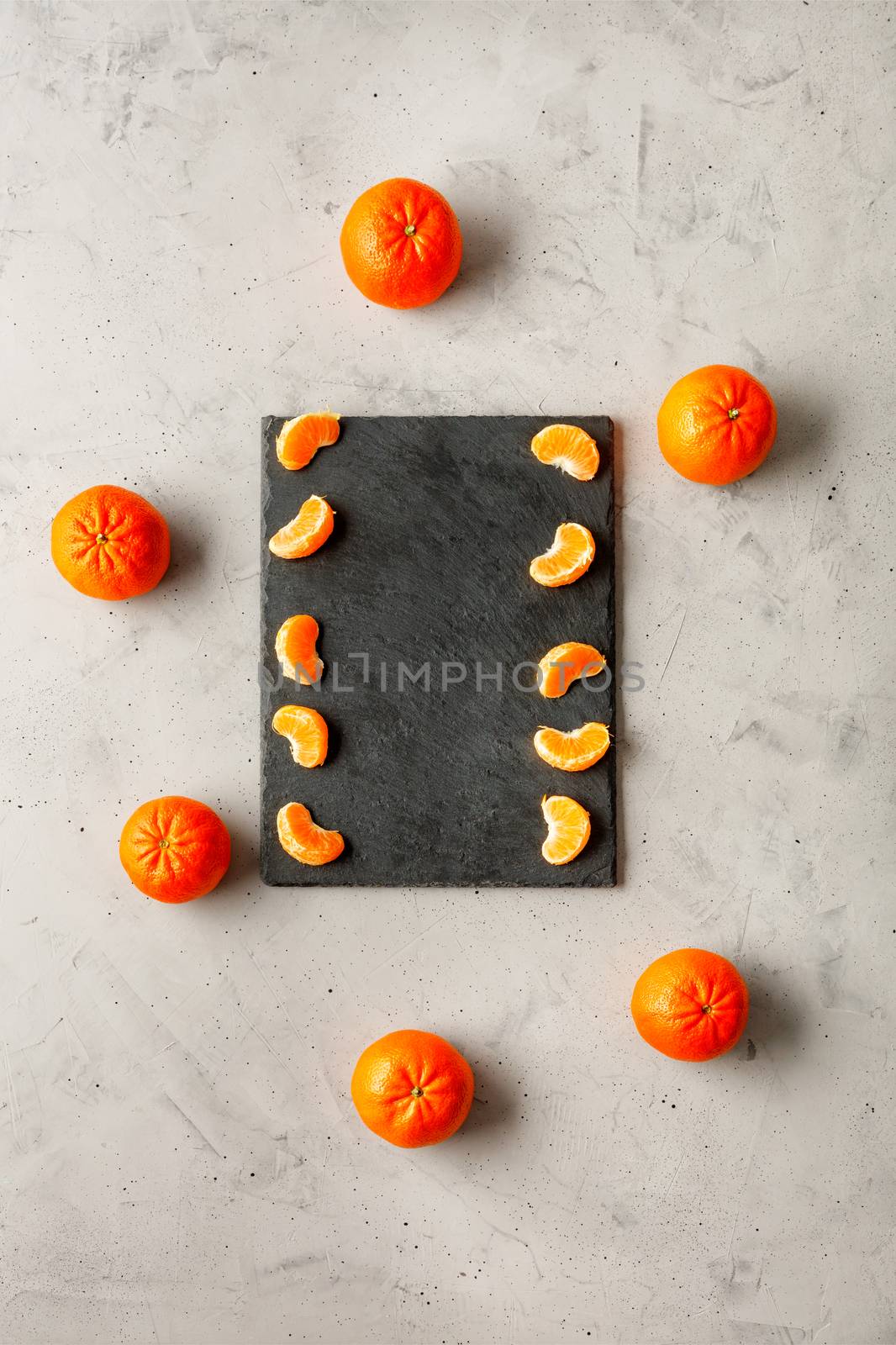 Pieces of tangerine, bright and juicy, lie on a gray slate surrounded by whole orange tangerines located on a gray concrete background, copy space, vertical image.