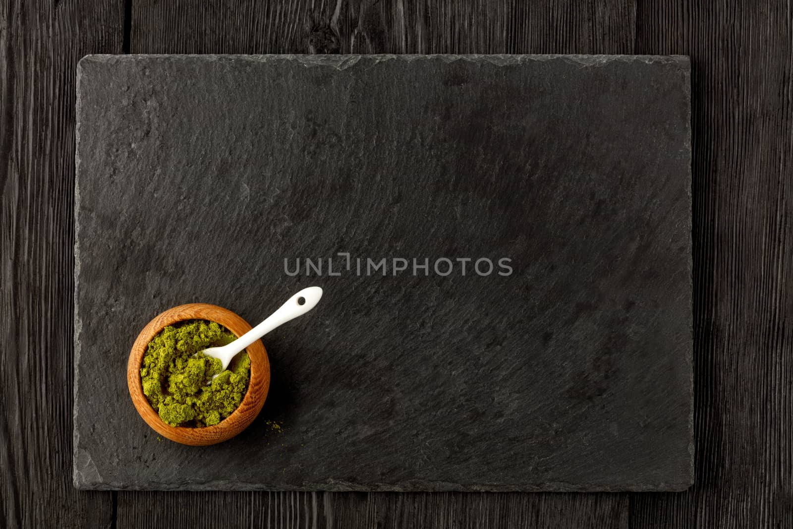 Matcha green tea powder in oak cup with white ceramic spoon with kitchen cutting board made of black slate, on wooden dark background, flat lay, copy space.