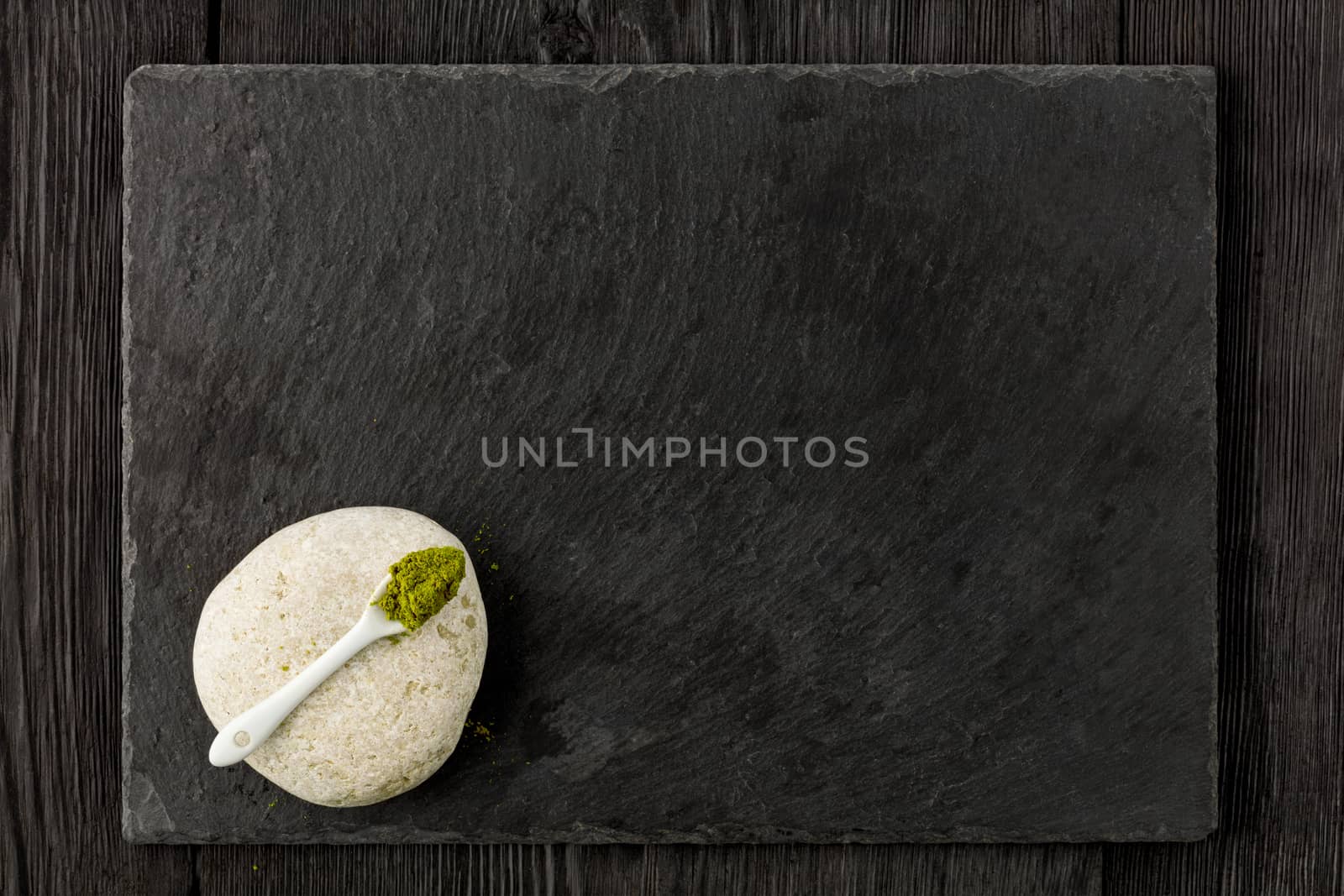 Matcha green tea powder in white ceramic spoon on white stone with kitchen cutting board made of black slate on wooden dark background, flat lay, copy space.