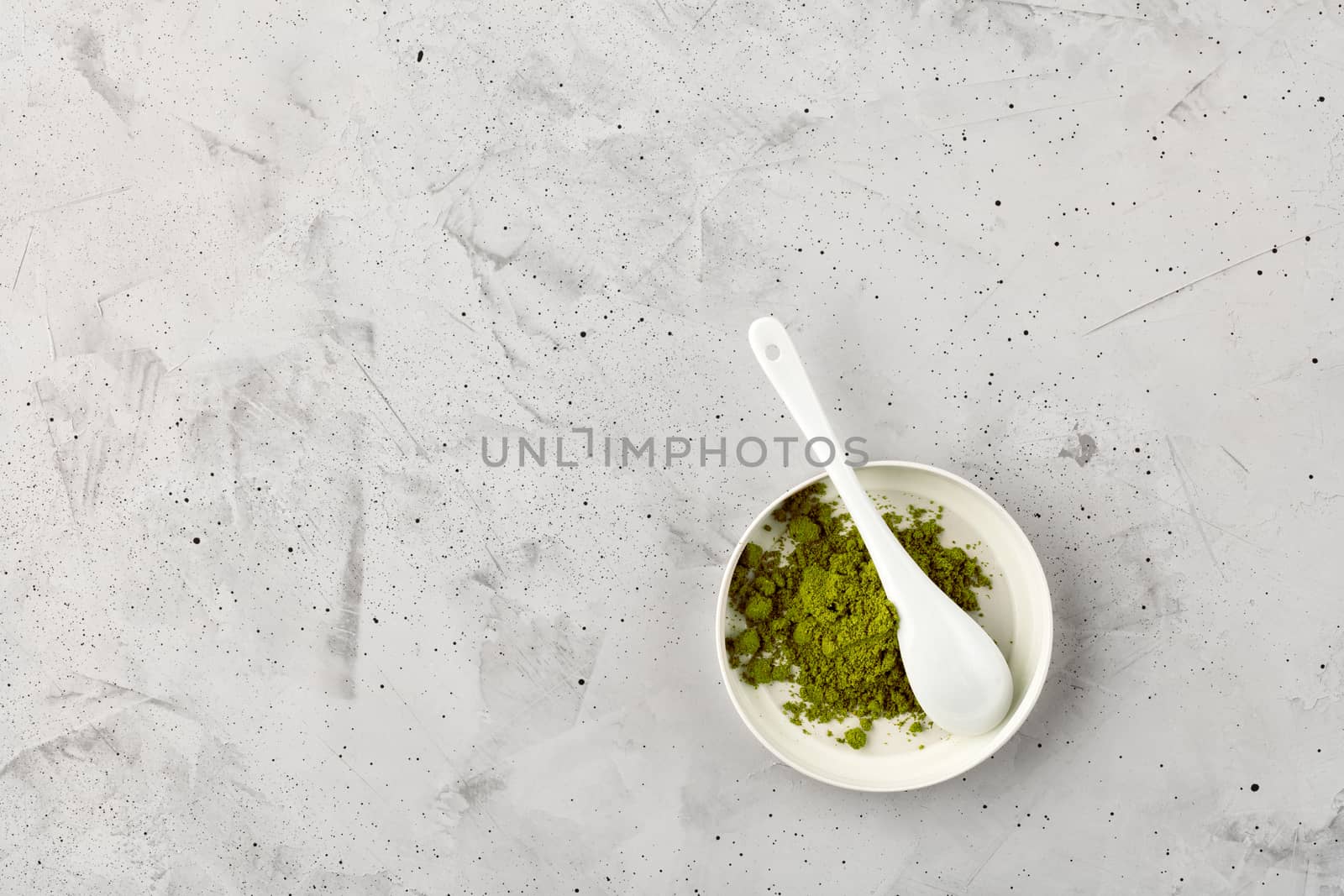 Matcha green tea powder in white ceramic cup with ceramic spoon on gray concrete background, flat lay, copy space.