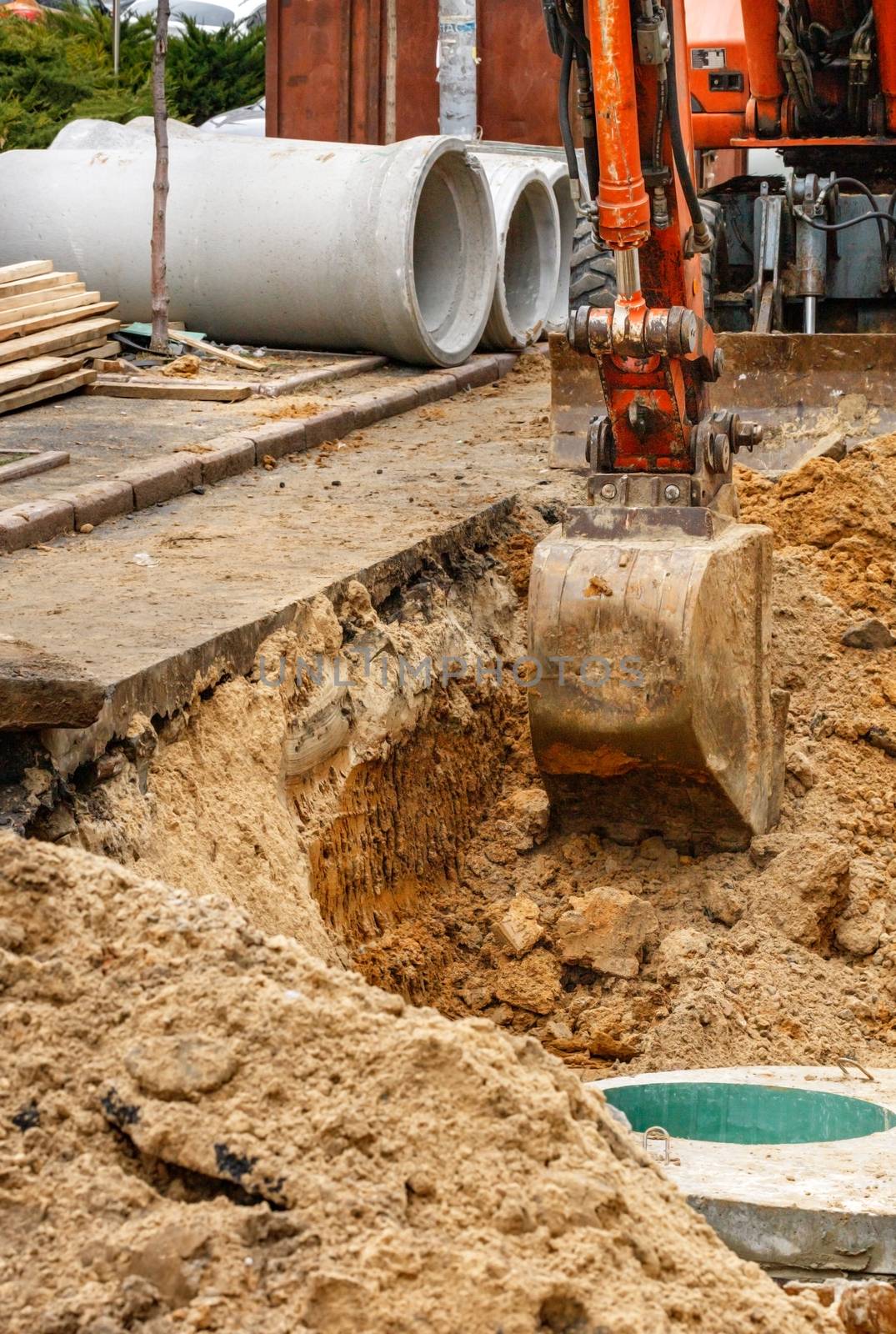 A bucket of a heavy construction excavator digs a trench, repairing a collector on a city street to replace old concrete blocks with new wells and sewer pipes.