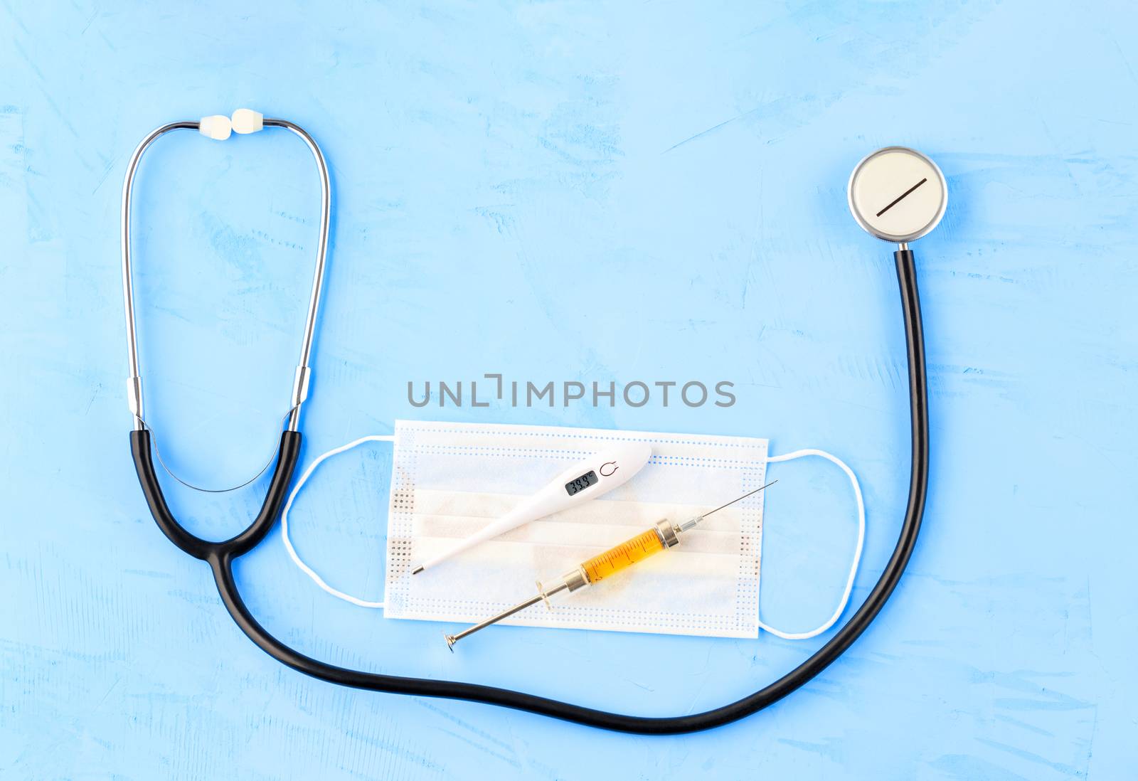 Stethoscope, medical mask, electronic thermometer and syringe with a yellow vaccine on a blue concrete background. The concept of protection against viruses, influenza and high body temperature. Copy space flat lay.