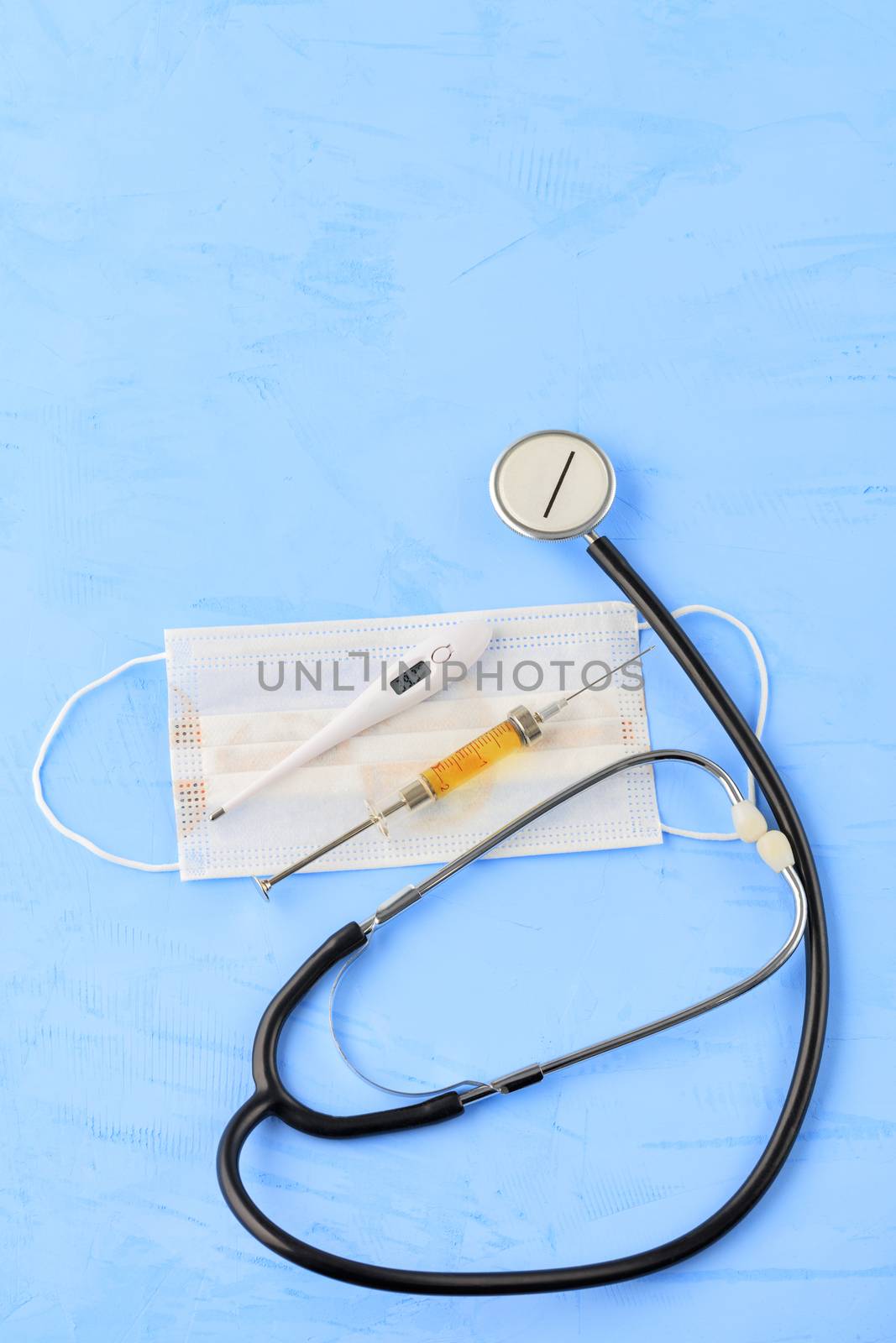 Stethoscope, medical mask, electronic thermometer and syringe with a yellow vaccine on a blue concrete background. The concept of protection against viruses, influenza and high body temperature. Copy space, vertical image.