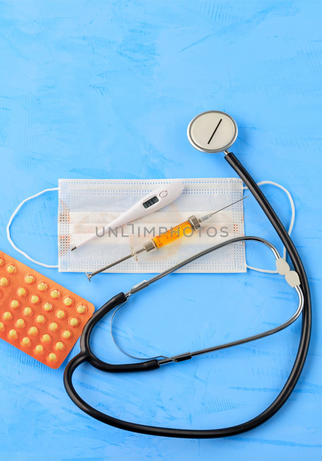 Stethoscope, medical mask, electronic thermometer, tablets and syringe with a yellow vaccine on a blue concrete background. The concept of protection against viruses, influenza and high body temperature. Copy space, vertical image.