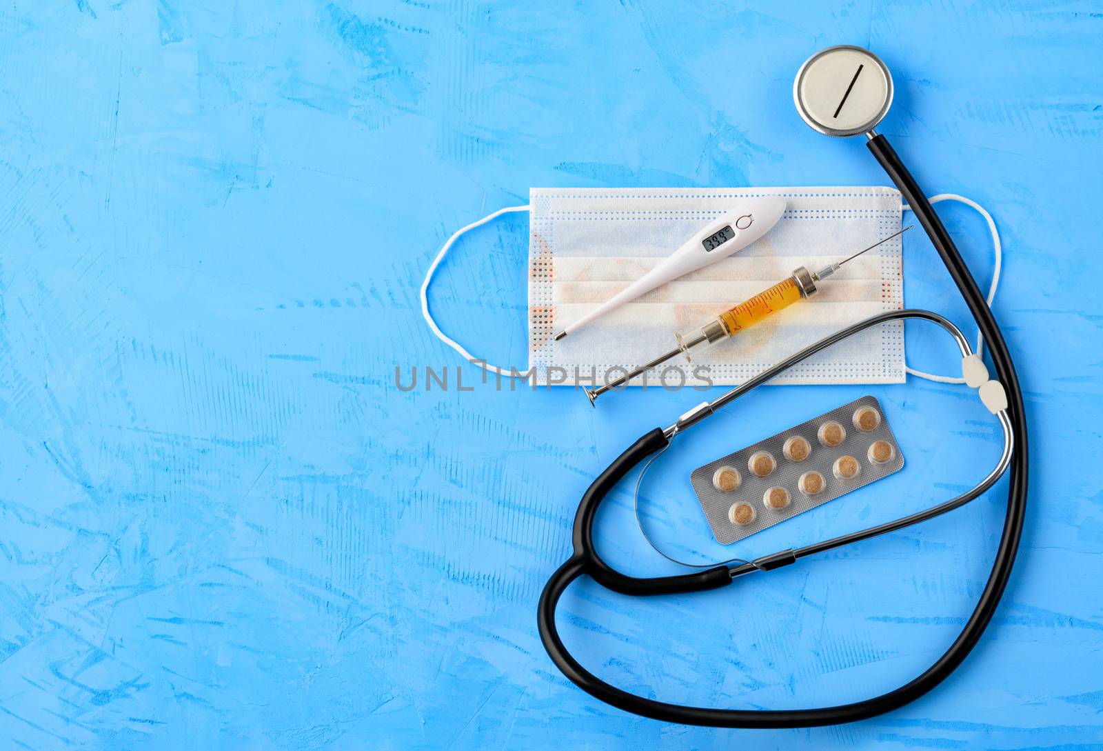 Stethoscope, medical mask, electronic thermometer and syringe with a yellow vaccine on a blue concrete background. The concept of protection against viruses, influenza and high body temperature. Copy space, top view.
