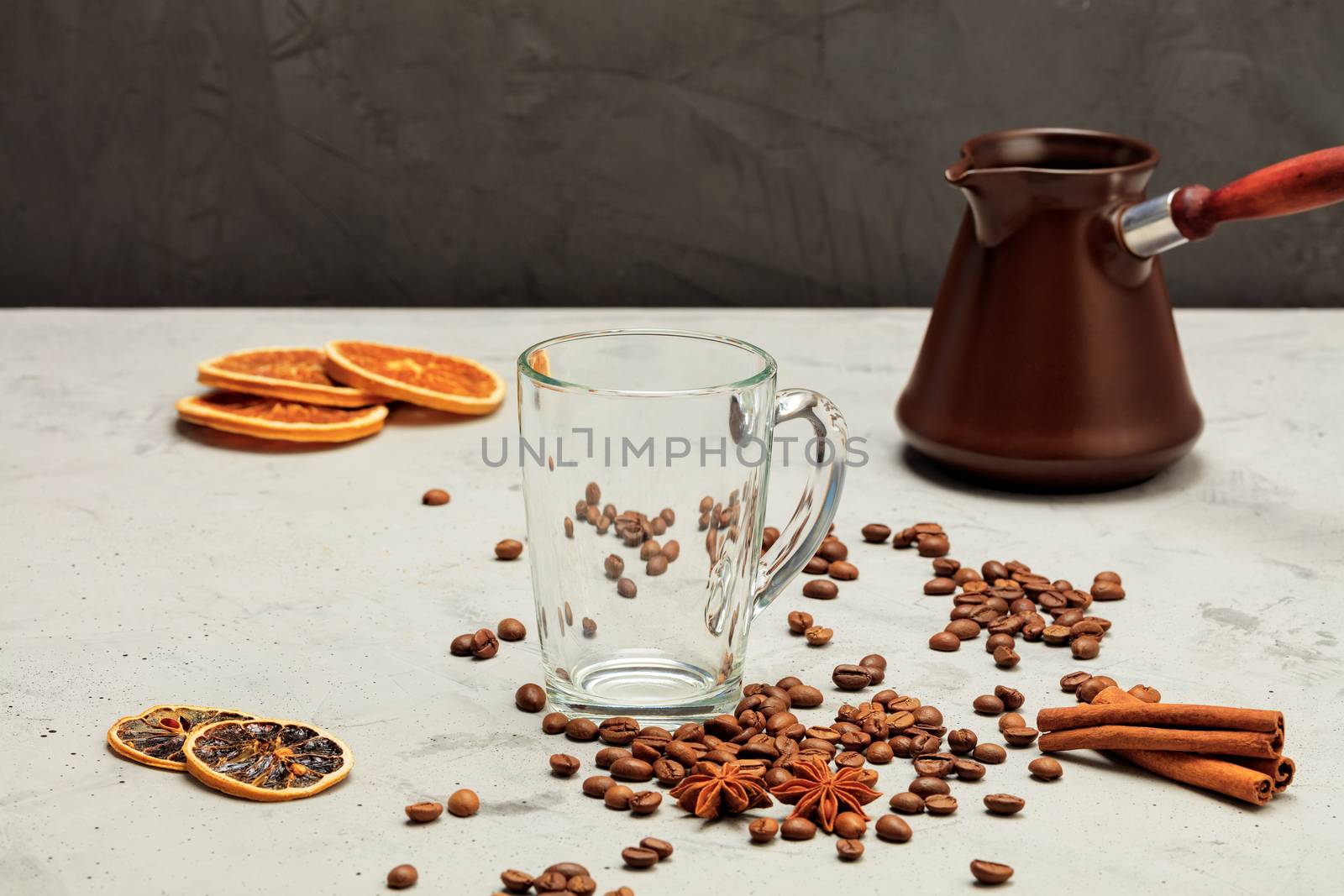 A transparent glass cup is surrounded by coffee beans, dried orange and lemon, anise stars and cinnamon sticks on a background of gray concrete and a coffee cezve in blur.